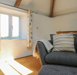 A sitting area with a grey armchair a patterned cushion a window with curtains and wooden floor at Sunbury Barn in St Agnes