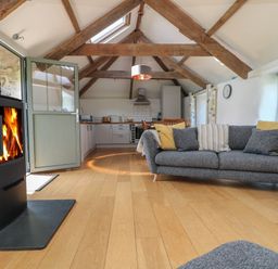 A living room with a fireplace a gray sofa with cushions wooden beams and an open door at Sunbury Barn in St Agnes