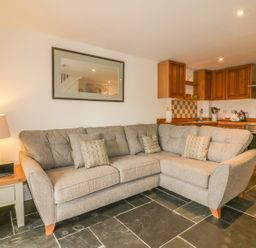 A living room with a gray sectional sofa and a lamp on a side table in an open kitchen with wooden cabinets and tiled backsplash at Crift Farm Cottage Lanlivery near Lostwithiel