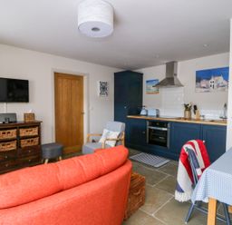 A living area with an orange sofa a grey armchair a wooden cabinet with baskets a wall-mounted TV and a kitchen with blue cabinets at Wallerthwaite Barn Cottage in Markington