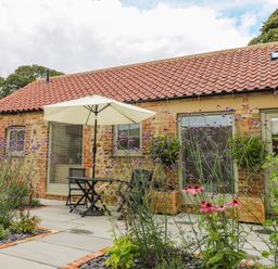A patio area with a table umbrella and chairs outside a brick cottage with flowers in the garden at Wallerthwaite Barn Cottage in Markington