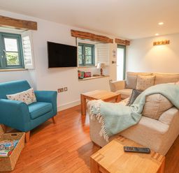 A living room with a beige sofa a blue armchair a wooden coffee table a wall mounted television and windows with shutters at Lantern Cottage in Padstow
