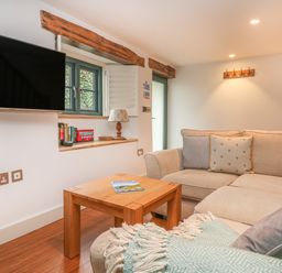 A living room with a beige sofa, wooden coffee table, wall-mounted TV, and a window with wooden beams at Lantern Cottage in Padstow