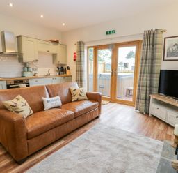 A living room with a brown leather sofa kitchen area and tv near glass doors with curtains at Wagtail Nest Northfield Cottage in Scalby