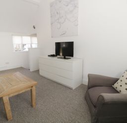 A living room with a wooden coffee table a gray armchair with a patterned pillow a white dresser with a television on top and three windows with blinds at Orchard Cottage in Shaftesbury