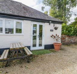An outdoor area with a wooden picnic table beside a white building with double glass doors and a potted plant at Orchard Cottage in Shaftesbury