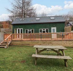 A backyard with a wooden picnic table a deck with railings and steps and a green wooden house with windows and a door at The Hen Pen in Star near Cenarth