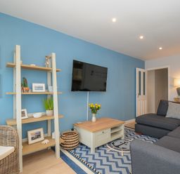 A living room with a blue wall a wall-mounted television a wooden shelf a coffee table and a dark grey sofa at Coastal Haven in Crackington near Crackington Haven