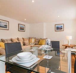 A dining area with a glass table set with plates and glasses and a living space with a sofa and framed pictures on the wall at The Crows Nest in Mevagissey