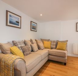 A beige sectional sofa with yellow and plaid cushions and a throw in a room with wooden floor and framed pictures on the wall at The Crows Nest in Mevagissey