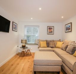 A living room with a beige sectional sofa and ottoman a wall-mounted tv and framed pictures at Captains Cabin in Mevagissey