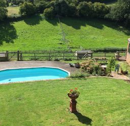 A backyard with a swimming pool surrounded by wooden decking a lawn with a flower pot a garden area with plants a picnic table and a brick building at The Lookout in Llangrove