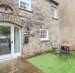 A stone cottage exterior with a white door a small window a black mailbox a wall lamp and a metal table with four chairs on artificial grass at Linden Cottage Ravenstonedale Nr Kirby Stephen