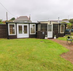 A garden with a patio table and chairs in front of a wooden building with white doors and windows at The Lodge in Rosemarket