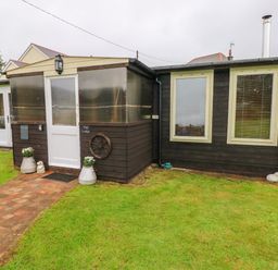 A dark wooden building with two windows and a white door surrounded by grass at The Lodge in Rosemarket