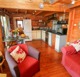 A wooden kitchen and living area with a fridge, stove, armchair, couch, and table with flowers at The Lodge in Rosemarket