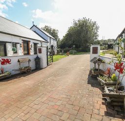 A paved driveway with white stone buildings on each side and plants in wooden containers at Cowslip Cottage in Rosemarket