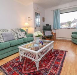 A living room with green sofas a wicker coffee table on a red patterned rug and a window with blue curtains at Phenests in Falmouth