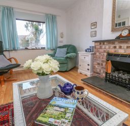 A living room with a glass coffee table with flowers cups and a magazine in front of a brick fireplace with a clock and two chairs at Phenests in Falmouth