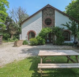 A house with arched windows surrounded by trees and plants a wooden picnic table on grass and a gravel driveway at Beggars Roost in Penhallow
