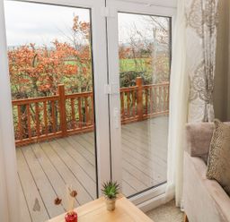 A corner of a living room with a beige sofa and pillows next to glass doors opening to a wooden deck with a railing and a view of trees and green fields at Avonal Lodge in Dunning