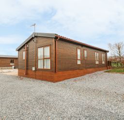 A wooden cabin on a gravel driveway with trees in the background at Avonal Lodge 24 in Dunning