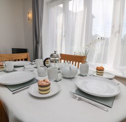 A dining table set with white plates, cups, and a teapot near a window with sheer curtains at Admiral's Quarter Apartment 4 in Weymouth