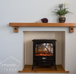 A small black electric stove heater in a recessed space with a wooden shelf above holding a vase with flowers and a small knitted item at Ammonite Cottage in Fortuneswell Portland