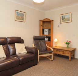 A living room with a leather sofa two cushions a wooden chair a wooden corner cabinet a wooden table with lamp and flowers and framed pictures on walls at Anvil House in Brewers Quay Harbour