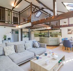 An open living space with a gray sectional sofa coffee table kitchen area and dining table under wooden loft beams at Barley Mews Brewers Quay Harbour