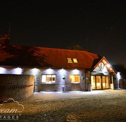 A brick cottage with exterior lights and a thatched roof at night with a gravel driveway at Baytree Lodge in Osmington