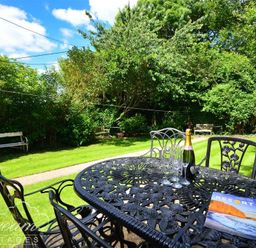 An outdoor garden area with a black metal table and chairs a bottle of champagne glasses and a travel magazine on the table benches and green trees in the background at Little Berwick Cottage Burton Bradstock