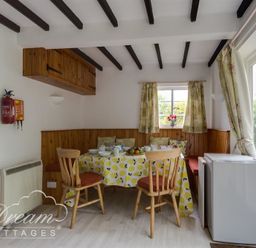A dining area with a table covered with a yellow-patterned tablecloth surrounded by wooden chairs and built-in bench seating at Little Berwick Cottage in Burton Bradstock