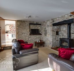 A living room with stone walls leather sofas red cushions a wood burning stove and a doorway leading to a kitchen at Blueberry Cottage in Sutton Poyntz
