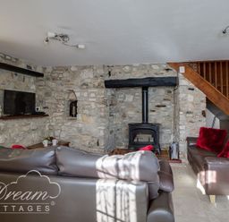 A sitting room with stone walls a wood burning stove two brown sofas with red cushions and wooden stairs at Blueberry Cottage in Sutton Poyntz