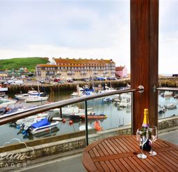 A balcony with a wooden table holding a bottle and two wine glasses overlooking a harbor with boats at The Boardwalk in West Bay