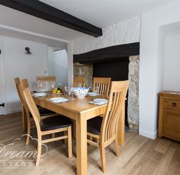 A dining room with a wooden table and six chairs near a stone fireplace and a wooden cabinet at Bramble Cottage in Burton Bradstock