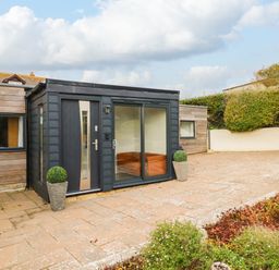 A patio area with potted plants and a wooden building with a black door and large glass window at Burton Bay in Burton Bradstock