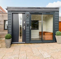A small modern building with a black door and glass sliding door between two potted plants on a paved patio at Burton Bay in Burton Bradstock