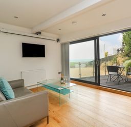 A living room with a gray sofa and glass coffee table facing a mounted TV and sliding glass doors at Burton Bay in Burton Bradstock