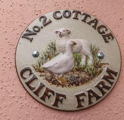 A circular sign on a pink textured wall with a lamb and a goose and text saying No 2 Cottage Cliff Farm