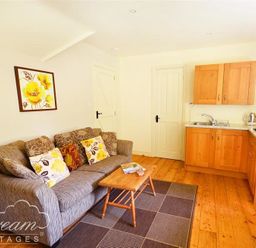 A living room with a sofa having floral cushions a wooden coffee table and a kitchen area with wooden cabinets at Coombe Orchard in Swanage