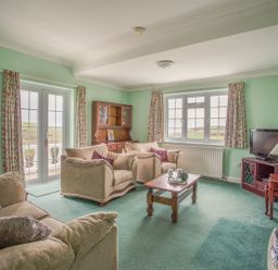 A living room with beige sofas a wooden coffee table a wooden cabinet a television and windows with floral curtains at The Doves in Burton Bradstock