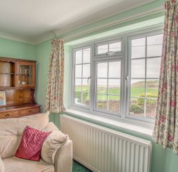 A sitting room with a beige sofa and floral curtains by a window with a countryside view at The Doves in Burton Bradstock