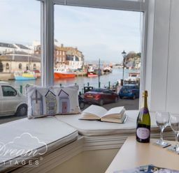 A window seat with a cushion and open book overlooking a harbor with boats and parked cars at Harbour Edge in Brewers Quay Harbour