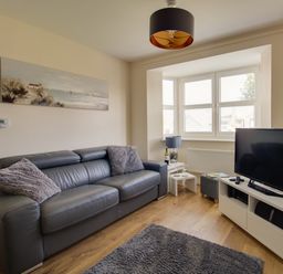 A living room with a grey leather couch and cushions a TV on a white stand and a bay window with blinds at The Ferns in Weymouth