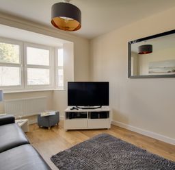 A living room with a black leather sofa a television on a white stand a grey rug and a window at The Ferns in Weymouth