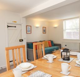 A dining area with wooden table and chairs set with white dishes and mugs next to a living area with a sofa and cushions at Firkin Cottage in Brewers Quay Harbour