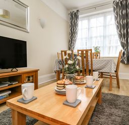 A living room with a wooden coffee table set with cups and muffins a wooden dining table with chairs against a window with curtains at Footprint in Brewers Quay Harbour