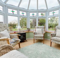 A conservatory with three armchairs a small stove a lamp and windows with stained glass at Grazeland Cottage Bridport